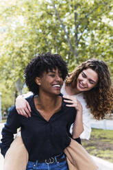 Cheerful woman giving piggyback ride to friend in park - PNAF05396