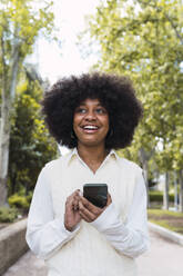 Happy Afro woman standing with smart phone in park - PNAF05402