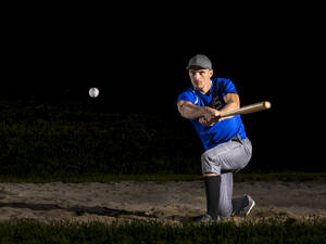 Young man hitting baseball with bat at night - STSF03744