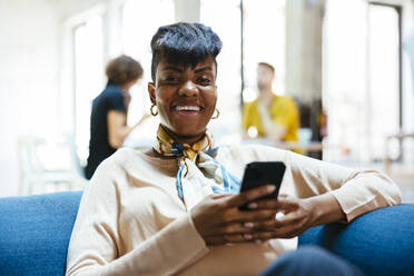 Glücklich lächelnde Geschäftsfrau sitzt mit Handy im Büro - EBSF03543