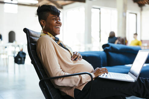 Glückliche Geschäftsfrau sitzt mit Laptop im Büro - EBSF03545