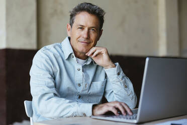Smiling mature businessman sitting with laptop at desk - EBSF03557