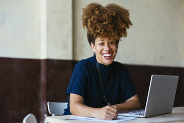 Glückliche Geschäftsfrau sitzt mit Laptop und Dokumenten am Schreibtisch - EBSF03559