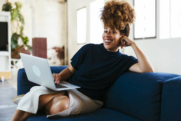 Happy businesswoman sitting with laptop on sofa in office - EBSF03575