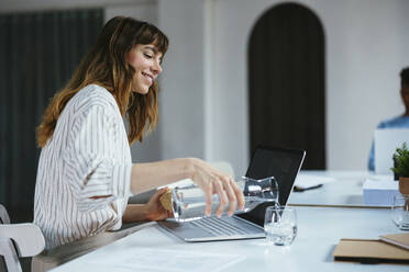 Glückliche Geschäftsfrau gießt am Schreibtisch Wasser in ein Glas - EBSF03595