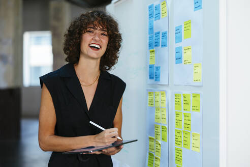 Happy young businesswoman with tablet PC and digitized pen standing by whiteboard in office - EBSF03617