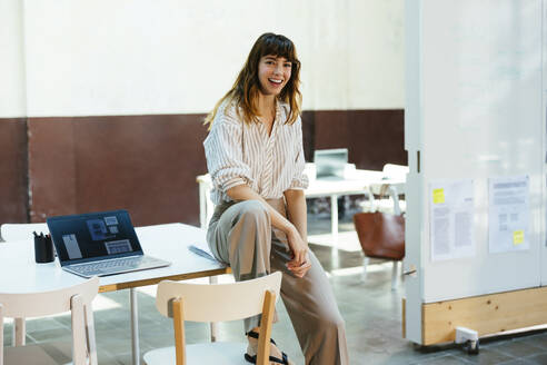 Lächelnde Geschäftsfrau sitzt auf dem Schreibtisch im Büro - EBSF03620