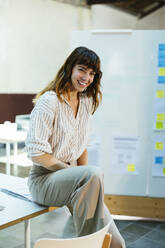 Happy businesswoman sitting on desk in office - EBSF03621