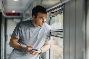 Mann hält Smartphone und lehnt sich an das Fenster eines Eisenbahnwaggons - VSNF01089