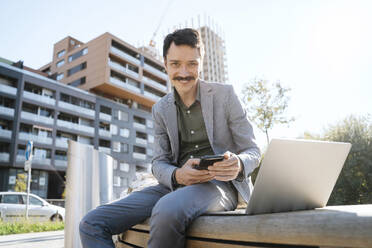 Lächelnder Geschäftsmann mit Smartphone und Laptop auf einer Bank im Büropark sitzend - NDEF00769