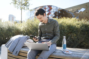 Businessman using smart phone and laptop sitting on bench at office park - NDEF00772