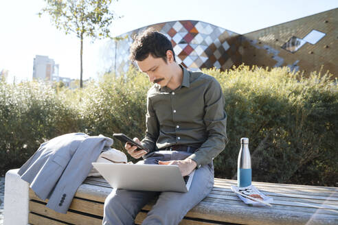 Geschäftsmann mit Smartphone und Laptop auf einer Bank im Büropark sitzend - NDEF00772