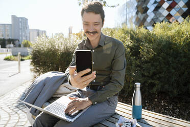 Lächelnder Geschäftsmann, der über sein Smartphone einen Videogespräch führt und mit seinem Laptop im Büropark sitzt - NDEF00773