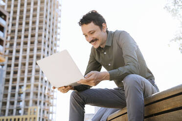 Smiling businessman holding laptop sitting at office park - NDEF00778