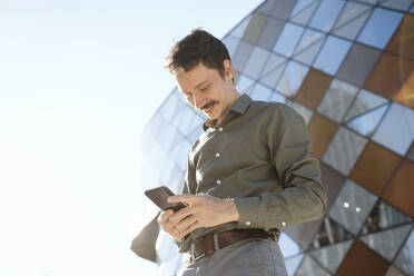 Lächelnder Geschäftsmann mit Smartphone vor einem modernen Bürogebäude - NDEF00782