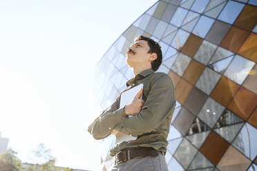 Thoughtful businessman holding laptop in front of modern office building - NDEF00784