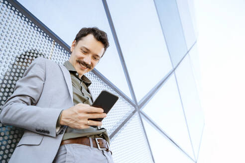 Lächelnder Geschäftsmann mit Smartphone vor einem modernen Glasgebäude - NDEF00794