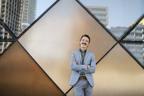 Confident businessman with arms crossed leaning on modern office building - NDEF00802