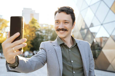 Ein fröhlicher Geschäftsmann mit einem eleganten Schnurrbart macht ein Selfie mit seinem Smartphone inmitten des Büroparks - NDEF00815