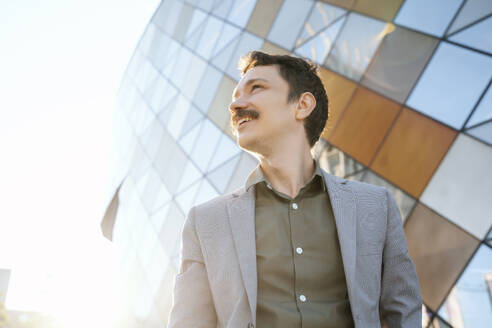 Businessman with mustache in suit standing by modern office building on sunny day - NDEF00816