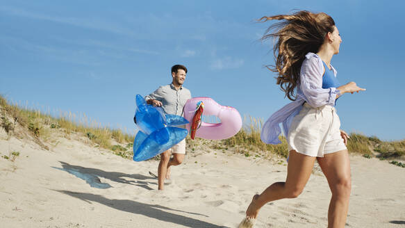 Glückliche Frau läuft mit einem Mann, der einen aufblasbaren Schwimmring und einen Spielzeug-Hai hält - ASGF03853