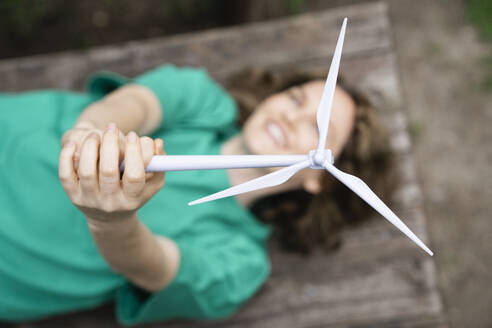 Playful woman holding wind turbine model lying on table at outdoors - EKGF00250