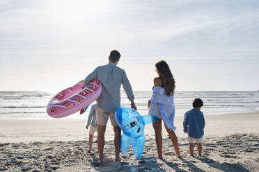 Familie mit aufblasbarem Schwimmring und Spielzeughai, die zusammen am Strand stehen - ASGF03857
