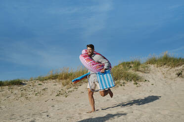 Happy man running with inflatable swim ring and folding chair at beach - ASGF03863