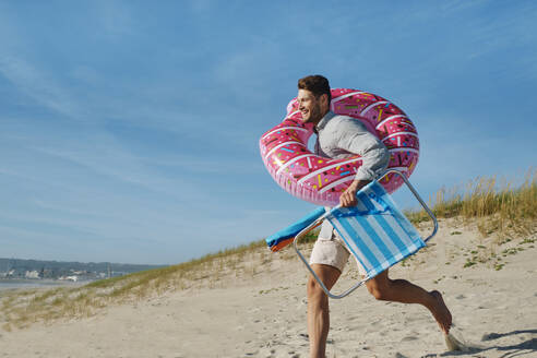 Happy man running with inflatable swim ring and folding chair at beach on sunny day - ASGF03864