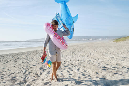 Happy man carrying inflatable swim ring and toy shark at beach - ASGF03868