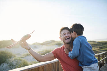 Happy man holding toy airplane and carrying son on sunny day - ASGF03875