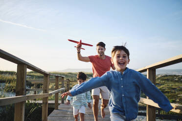 Father holding toy airplane with children running and enjoying on boardwalk - ASGF03878