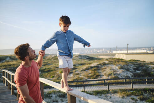 Glücklicher Vater mit Sohn, der auf einem Holzgeländer läuft - ASGF03883