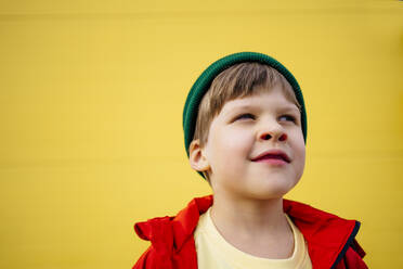 Nachdenklicher Junge mit roter Jacke und Strickmütze vor einer gelben Wand - MDOF01349