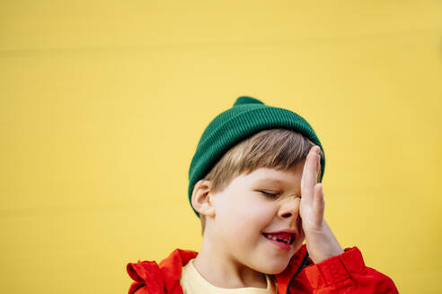 Playful boy wearing knit hat rubbing nose in front of yellow wall - MDOF01350