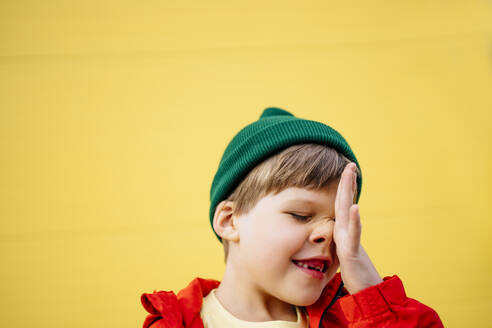 Verspielter Junge mit Strickmütze, der sich vor einer gelben Wand die Nase reibt - MDOF01350