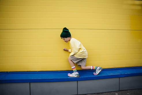Playful boy wearing sweatshirt crouching in front of yellow wall - MDOF01359