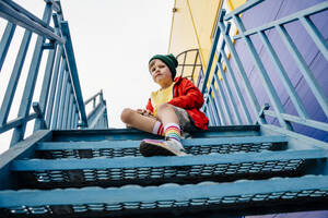 Junge in roter Jacke sitzt auf einer Metalltreppe am Gebäude - MDOF01363