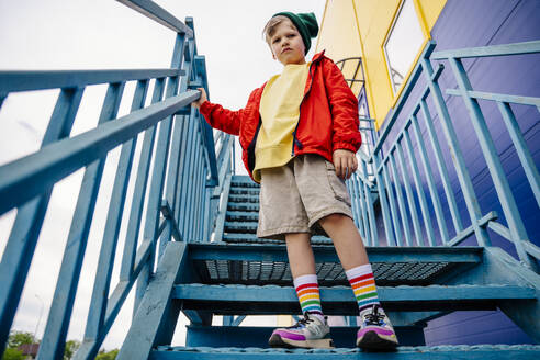 Boy in red jacket standing on metallic staircase by building - MDOF01366