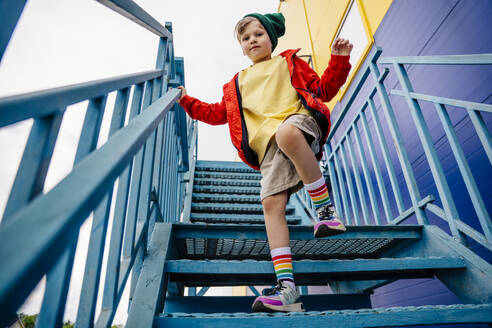 Boy in red jacket standing on one leg on metallic staircase by building - MDOF01367