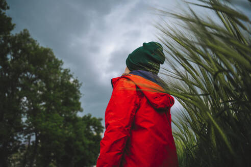 Junge in roter Jacke steht bei der Ernte auf einem Feld mit Wolken im Hintergrund - MDOF01385