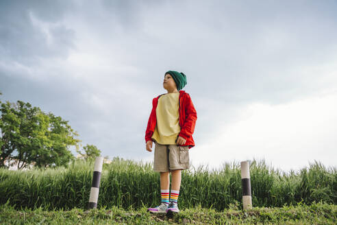 Boy in red jacket standing on barley field with cloudy sky - MDOF01388