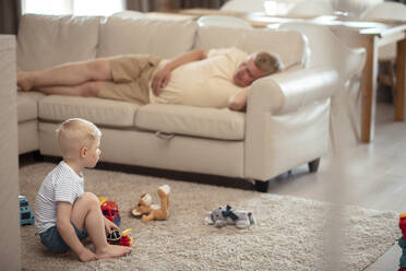 Sohn spielt mit Spielzeug und Vater schläft auf dem Sofa zu Hause - ADF00115