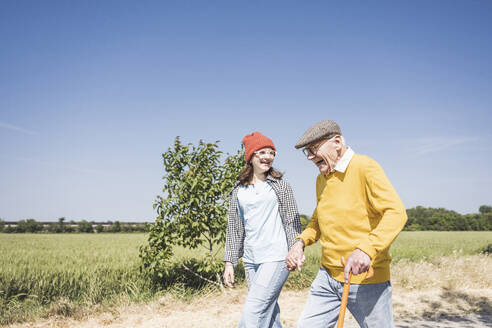 Happy senior man holding hands and walking with granddaughter on sunny day - UUF28943