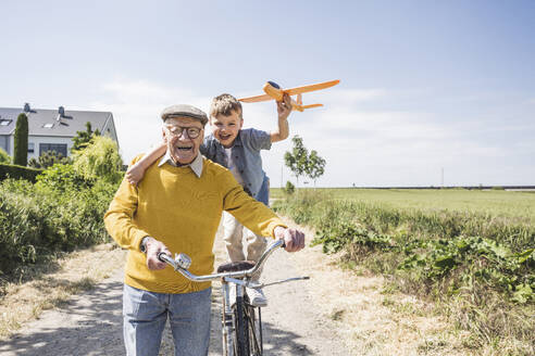 Happy boy holding toy airplane standing on bicycle and enjoying with grandfather - UUF28966