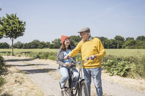 Glücklicher Großvater mit Enkelin auf dem Fahrrad sitzend auf einem Feld - UUF28971