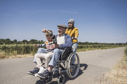 Happy girl with grandfather and grandson sitting in wheelchair on road - UUF29001