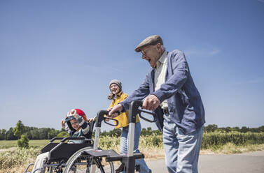 Glücklicher älterer Mann, der mit einer Gehhilfe spazieren geht, und Junge, der im Rollstuhl sitzt, an einem sonnigen Tag - UUF29004
