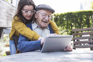 Girl teaching tablet PC to grandfather at table in backyard - UUF29014