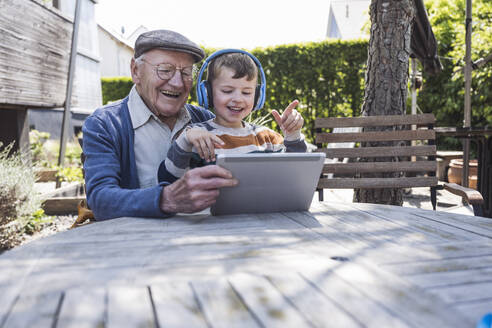 Glücklicher älterer Mann mit Enkel, der einen Tablet-PC am Tisch benutzt - UUF29020